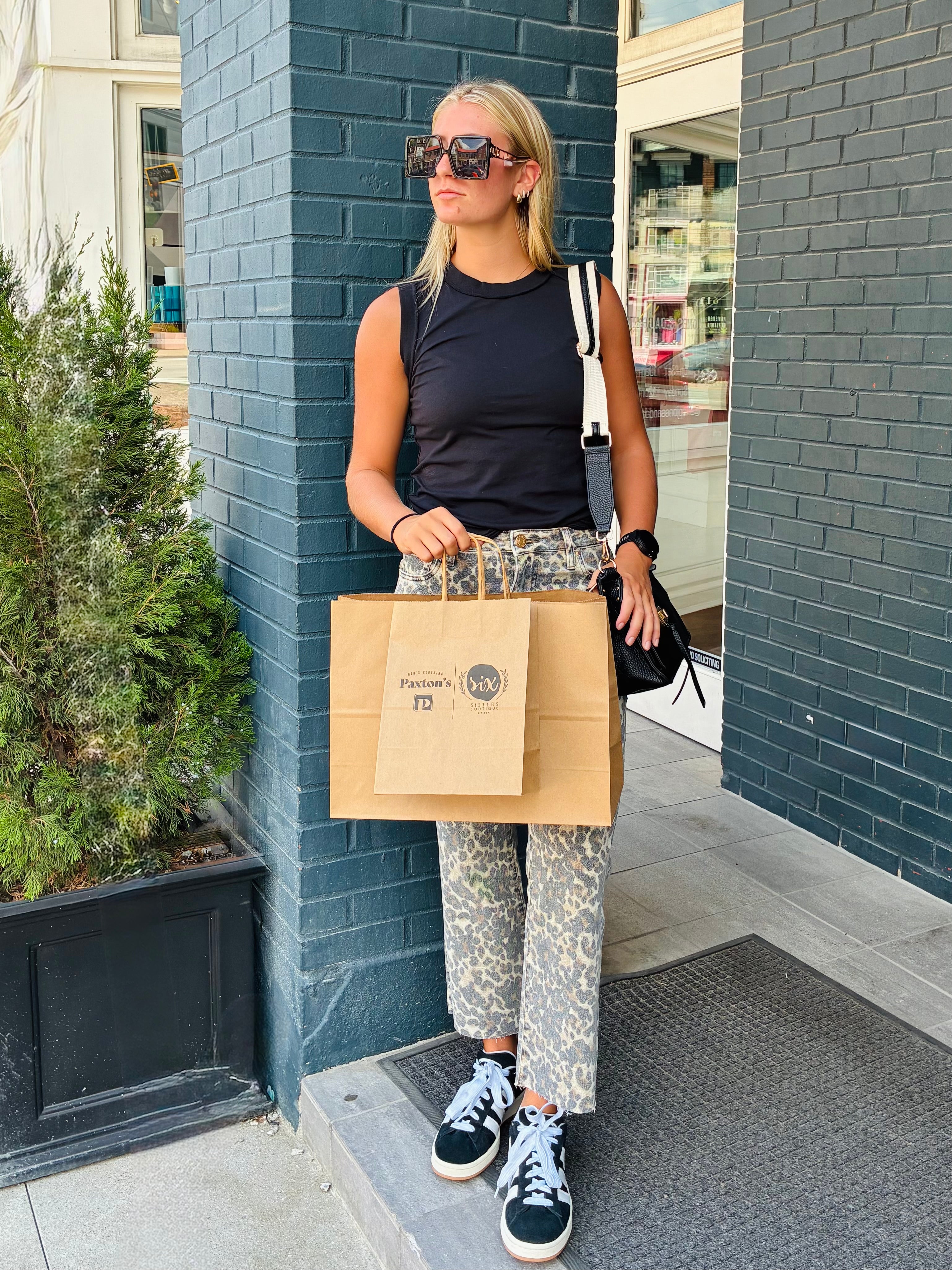 Woman holding a brown paper bag with a Six Sisters Boutique Store logo, standing against a blue brick wall in Nulu Louisville KY